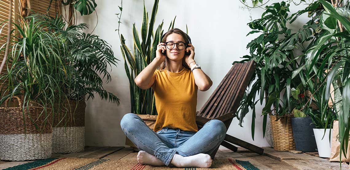 A woman sitting on the floor, surrounded by potted plants, listening to audio with headphones on.
