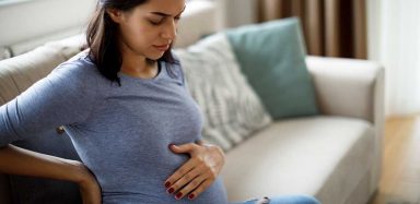 A pregnant mother sits on a couch while holding her back and her stomach.
