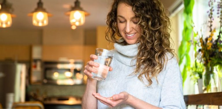 A woman with curly hair holding a glass of water and a supplement pill.